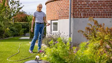 En kvinde i stribet t-shirt og jeans vander et blomsterbed med en haveslange foran en rund murstensbygning. Blomsterbedet er fyldt med farverige planter, og baggrunden viser en velplejet græsplæne og buske.