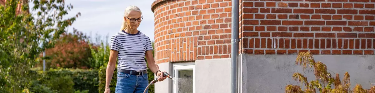 En kvinde i stribet t-shirt og jeans vander et blomsterbed med en haveslange foran en rund murstensbygning. Blomsterbedet er fyldt med farverige planter, og baggrunden viser en velplejet græsplæne og buske.