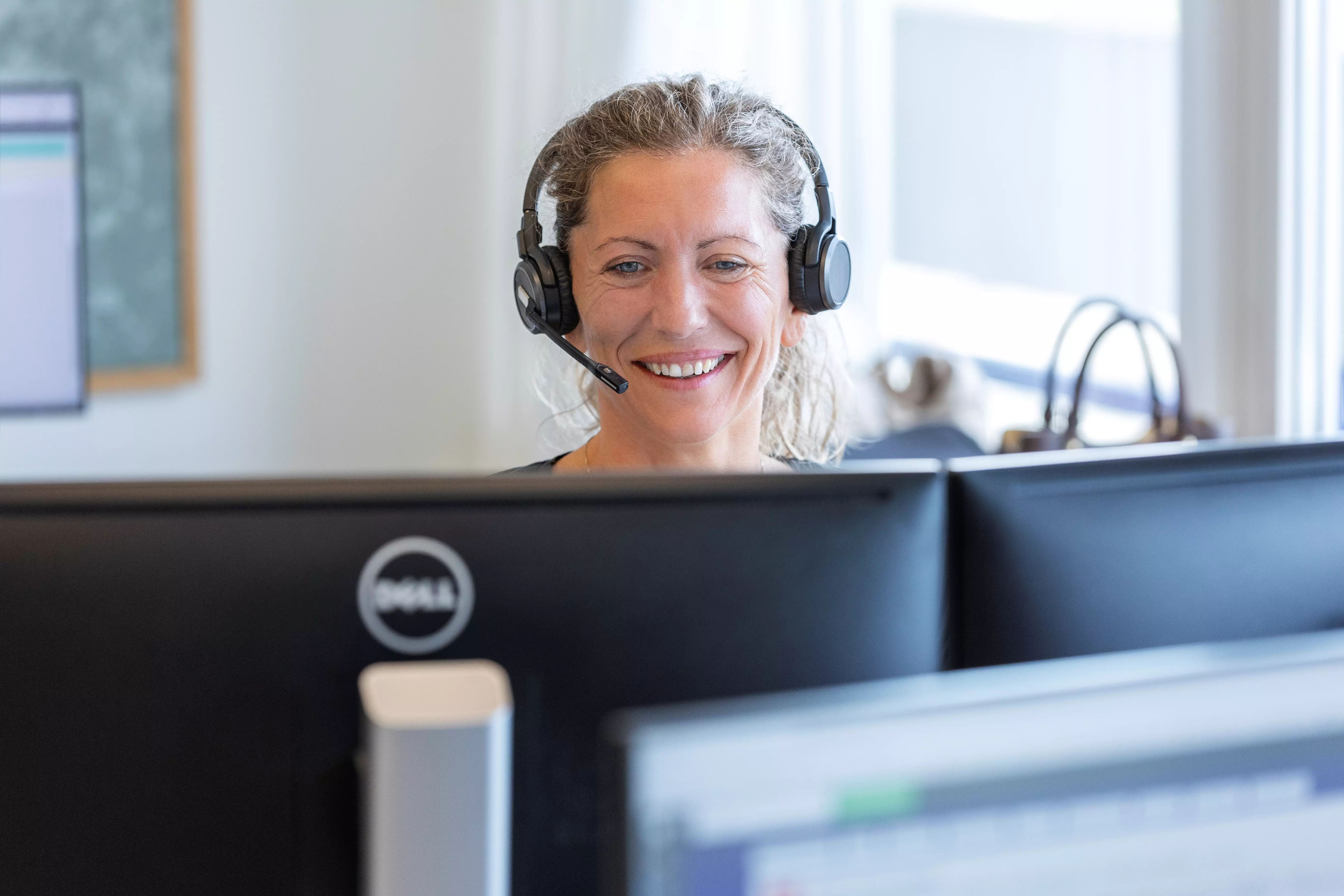 En kvinde snakker i telefon med headset og kigger smilende ind i sin computer