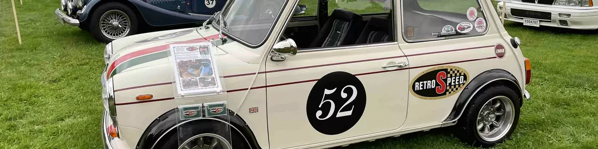 Billedet viser en veteranbil der holder på en græsplæne. I baggrunden ses flere biler.