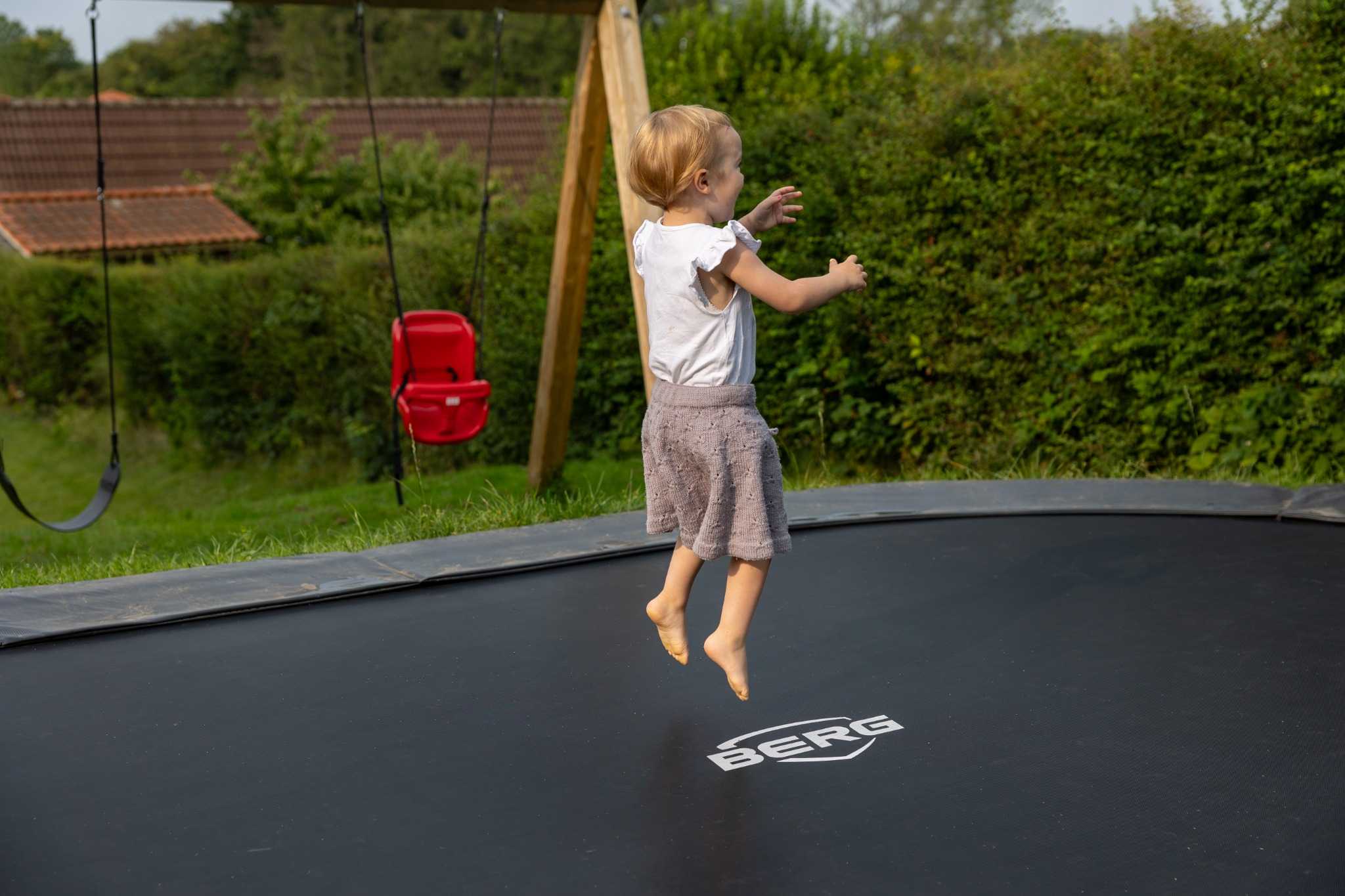 Et lille barn hopper på en sort trampolin i en have. I baggrunden ses en rød gynge og grønne buske.