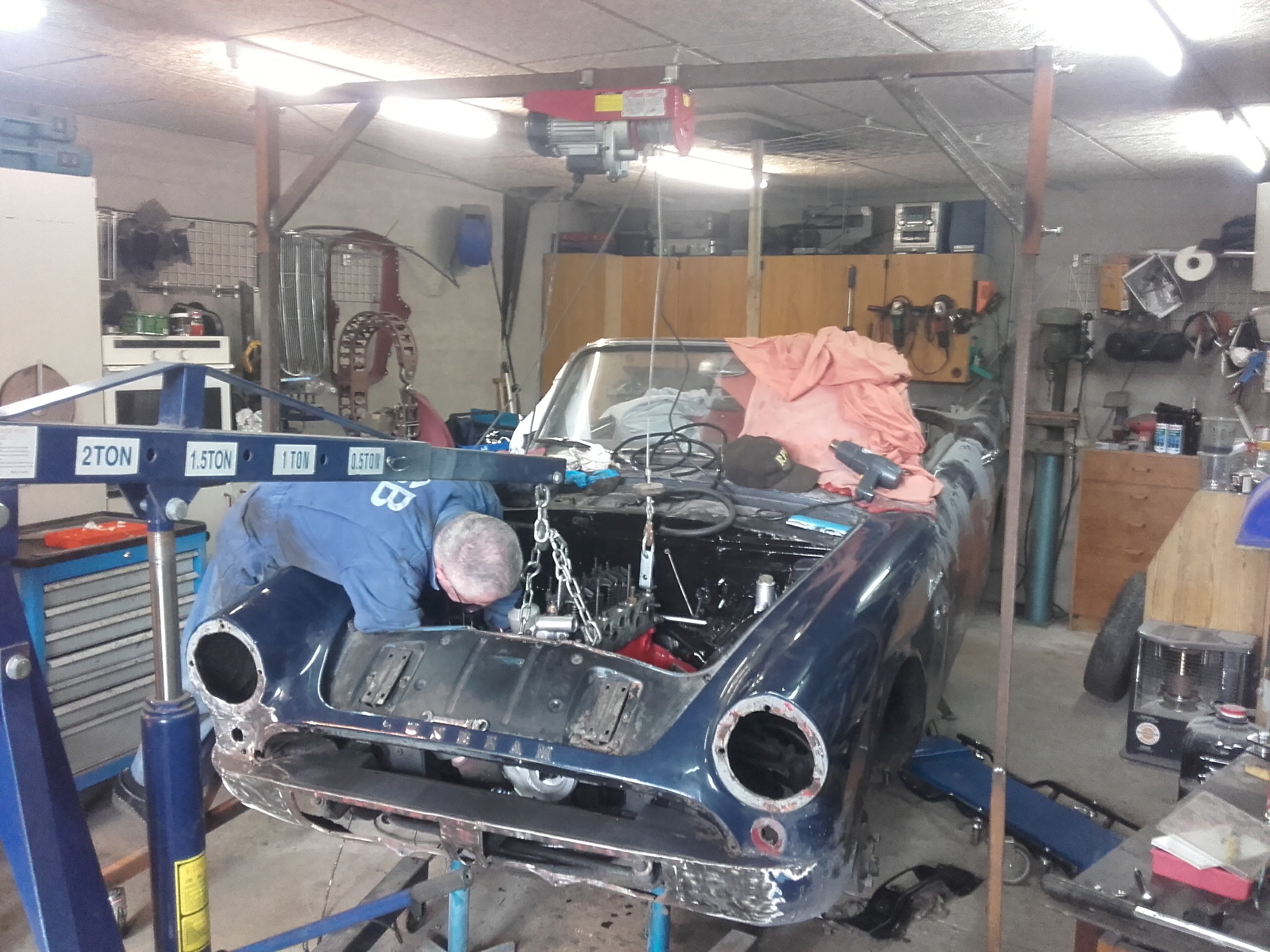 En klassisk bil holder på værksted, hvor en mekaniker er igang med at ordne noget på bilen. 