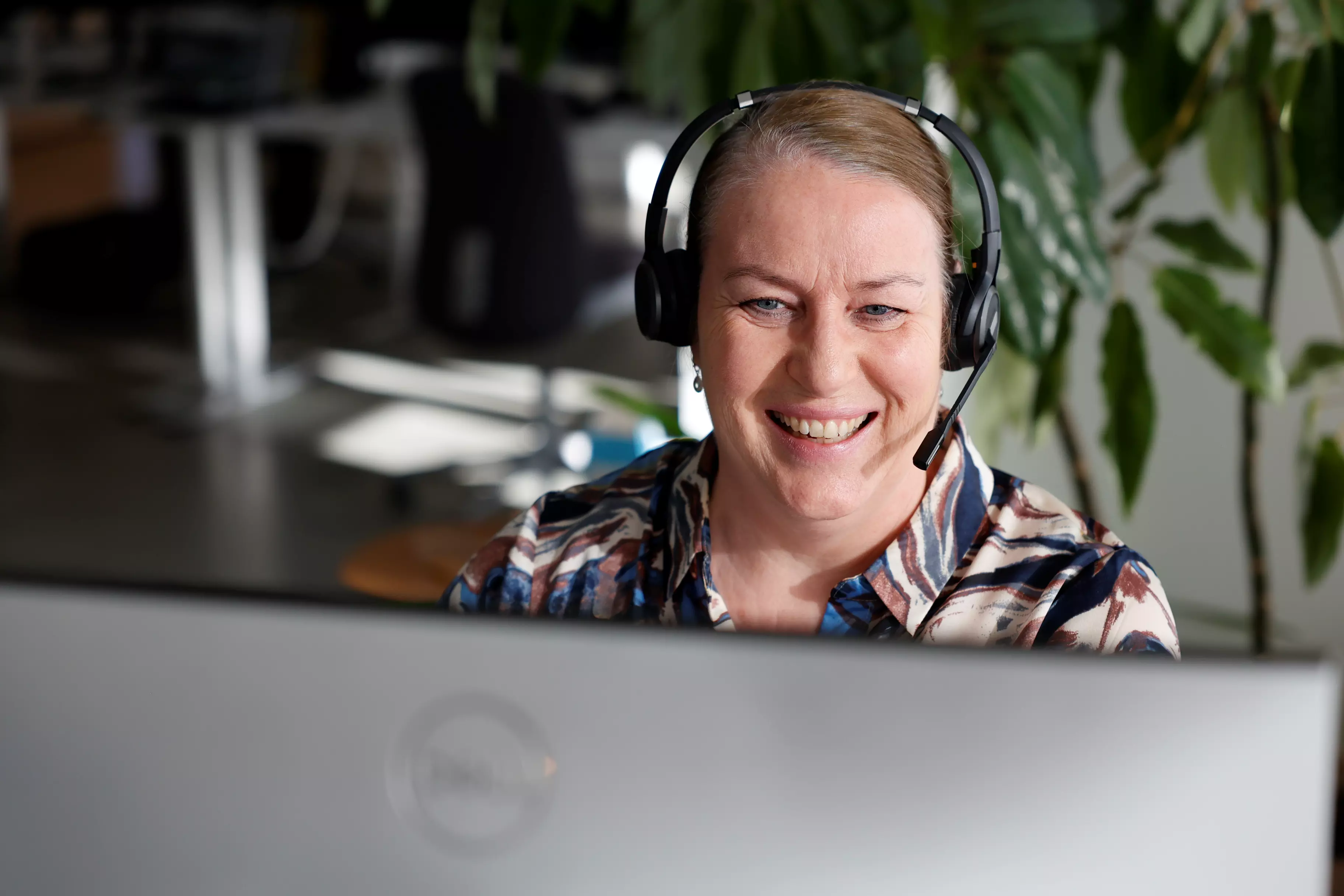 En kvinde sidder foran en computer og kigger ind i skærmen mens hun smiler. Hun har headset på hovedet. I baggrunden ses et kontormiljø.