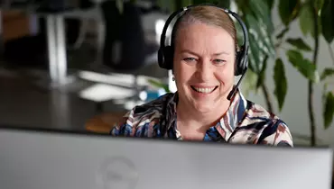 En kvinde sidder foran en computer og kigger ind i skærmen mens hun smiler. Hun har headset på hovedet. I baggrunden ses et kontormiljø.