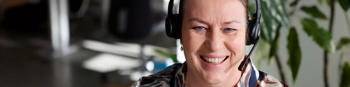 En kvinde sidder foran en computer og kigger ind i skærmen mens hun smiler. Hun har headset på hovedet. I baggrunden ses et kontormiljø.