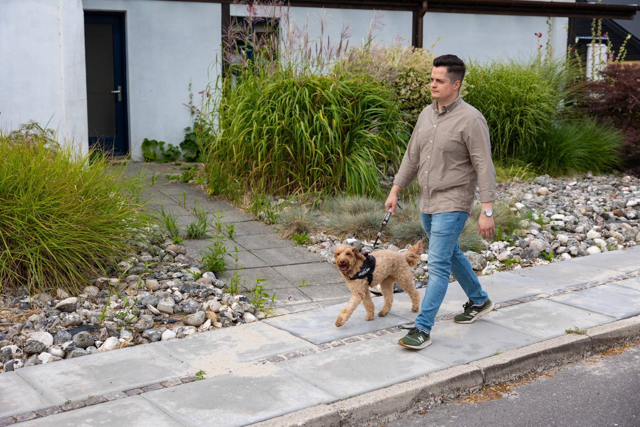 En mand går en tur med sin hund på en fortovssti foran et hus med en forhave fyldt med dekorative sten og grønne buske.
