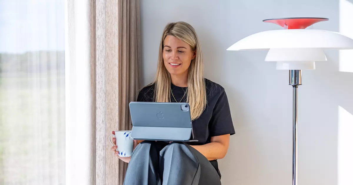 En kvinde sidder afslappet på en vindueskarm med benene trukket op, mens hun holder en tablet i den ene hånd og en kaffekop i den anden. Hun smiler og kigger på tabletten. Sollys strømmer ind gennem vinduet ved siden af hende.