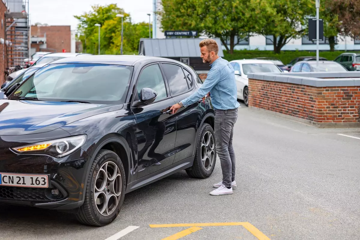 En mand iført denimskjorte og grå bukser åbner døren til en sort Alfa Romeo SUV parkeret foran en murstensbygning. Baggrunden viser træer og andre parkerede biler.
