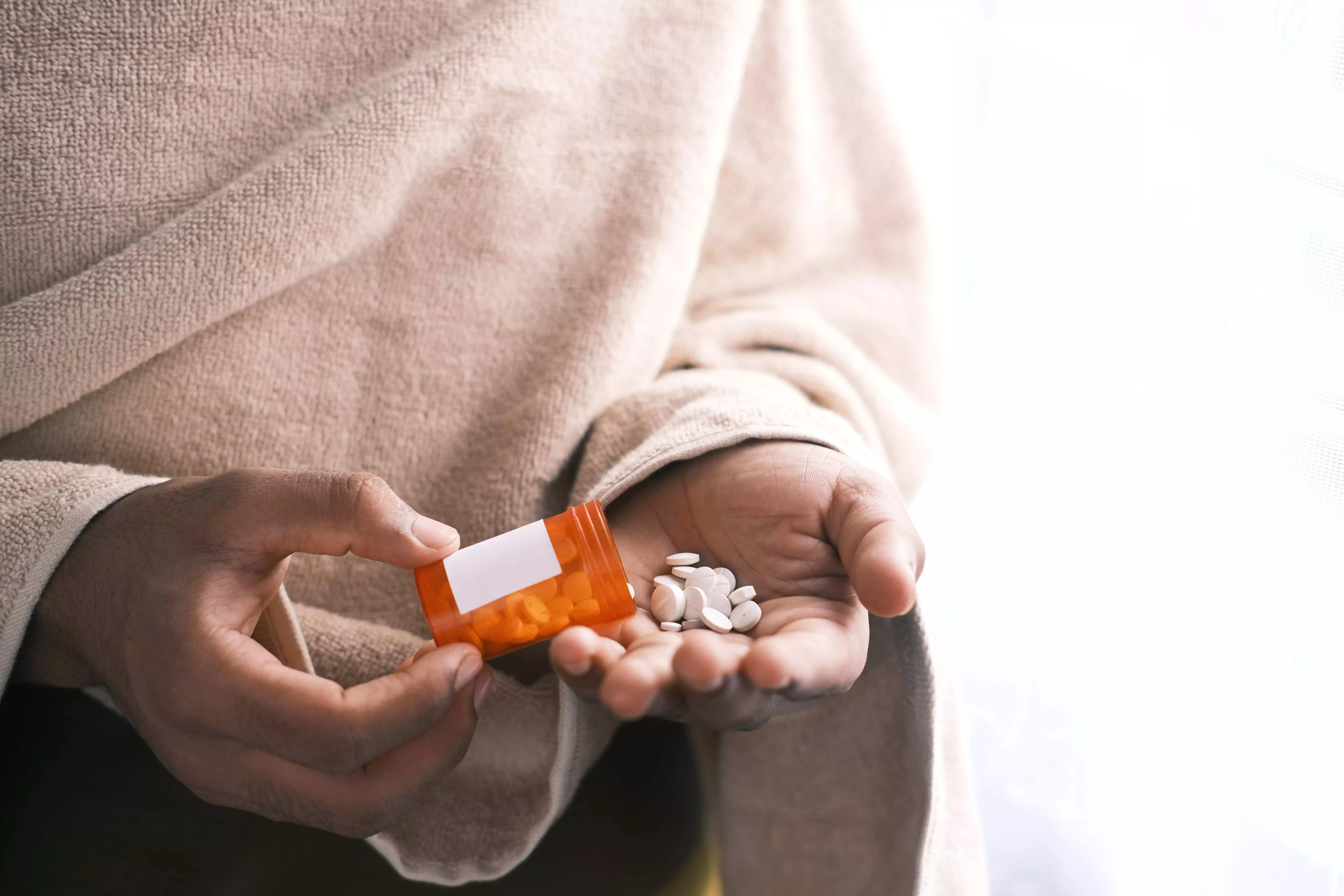 En person iført et beige tæppe hælder hvide piller ud i sin hånd fra en orange medicinflaske. Baggrunden er sløret og lys.