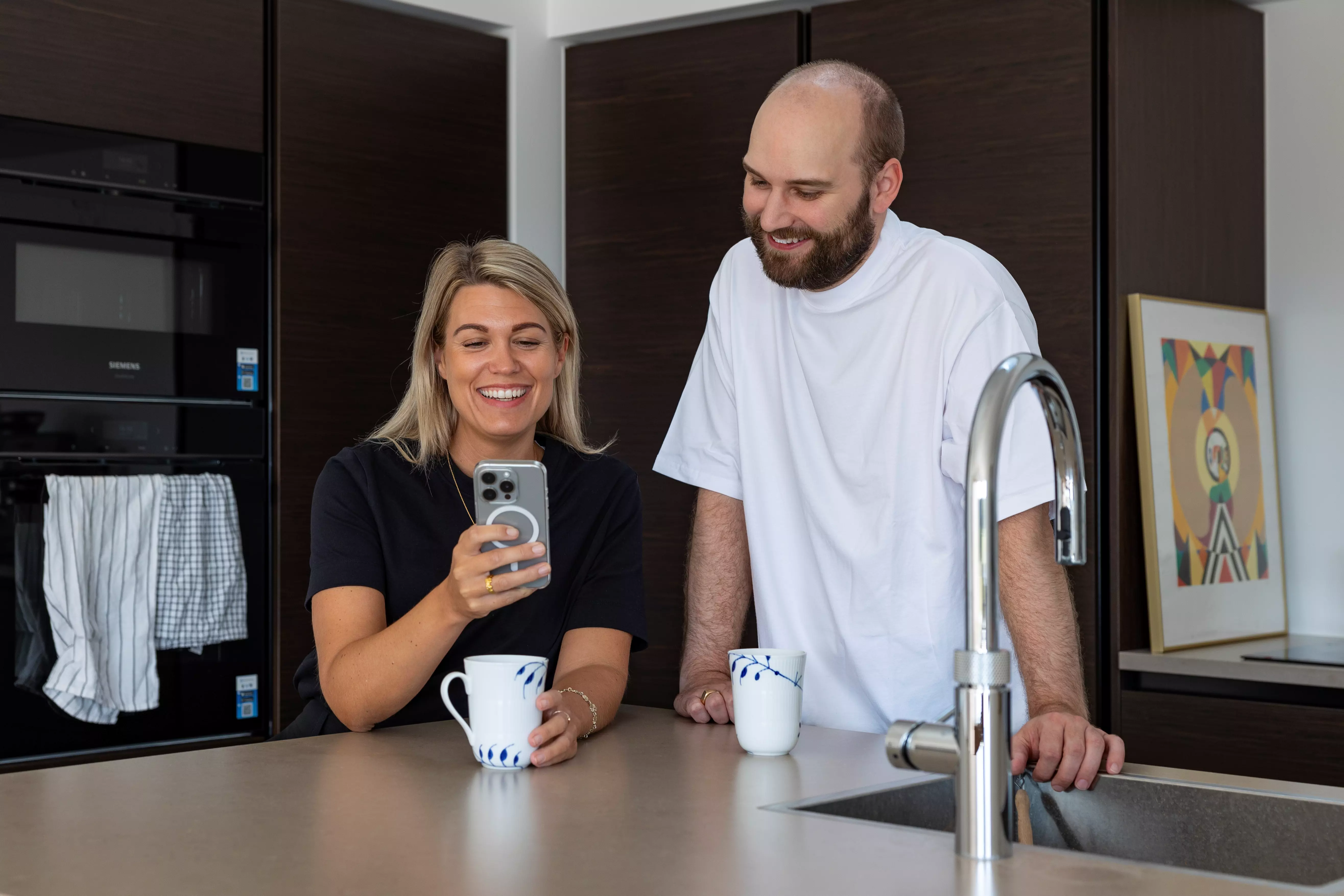 En mand og en kvinde står i et køkken og læner sig op af køkkenbordet. Kvinden holder sin telefon i hånden, mens manden ser til og smiler.