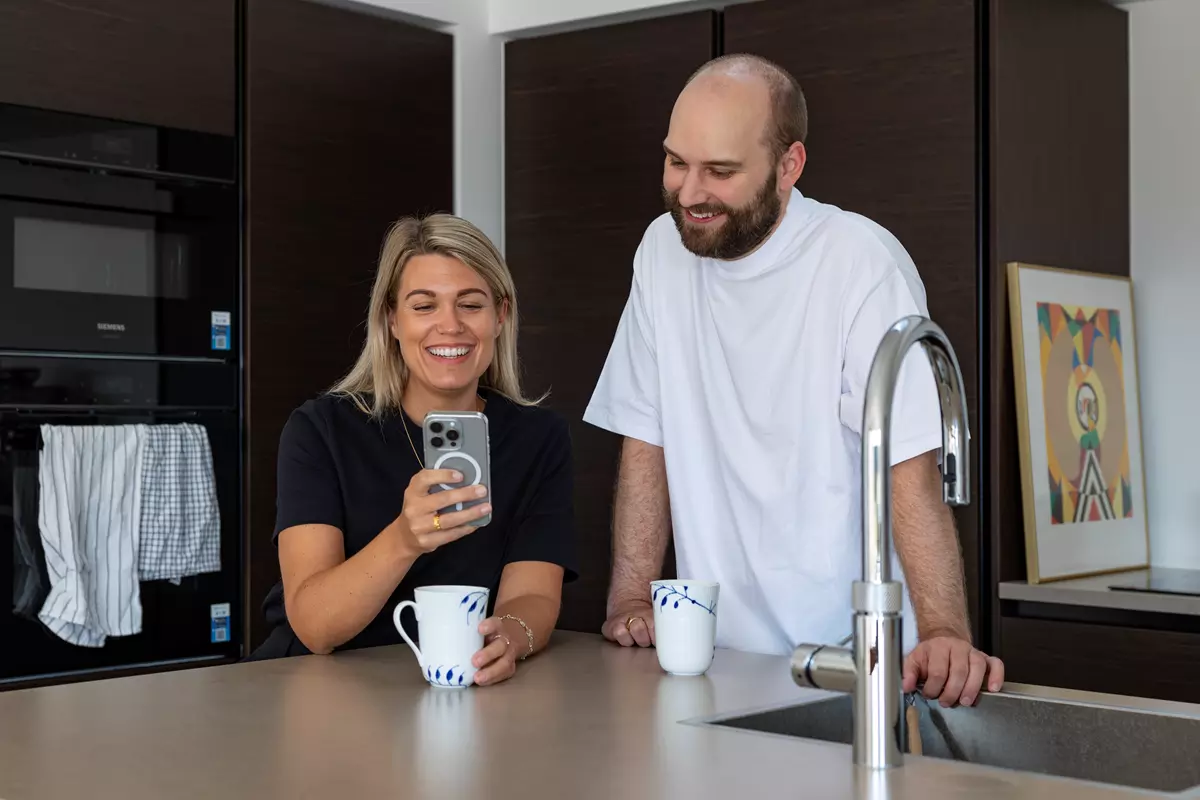 En mand og en kvinde står i et køkken og læner sig op af køkkenbordet. Kvinden holder sin telefon i hånden, mens manden ser til og smiler.