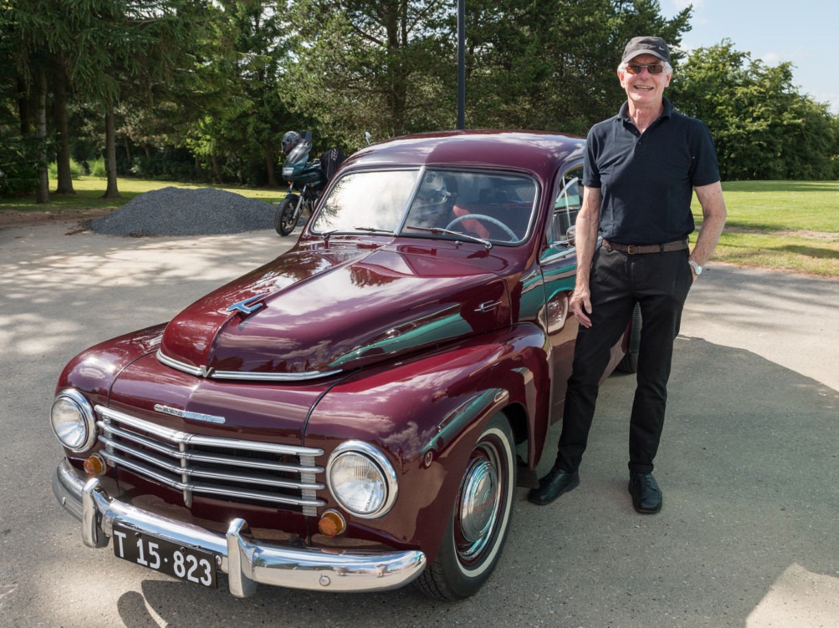 Billede af en rød klassisk bil. Der står en mand ved siden af bilen og smiler.
