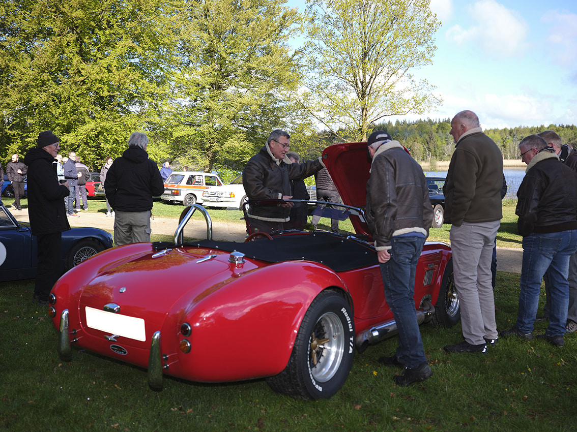 Klassisk bil, der er omringet af mennesker der kigger på bilen. Kølerhjelmen er åben og et par af menneskerne kigger ned.