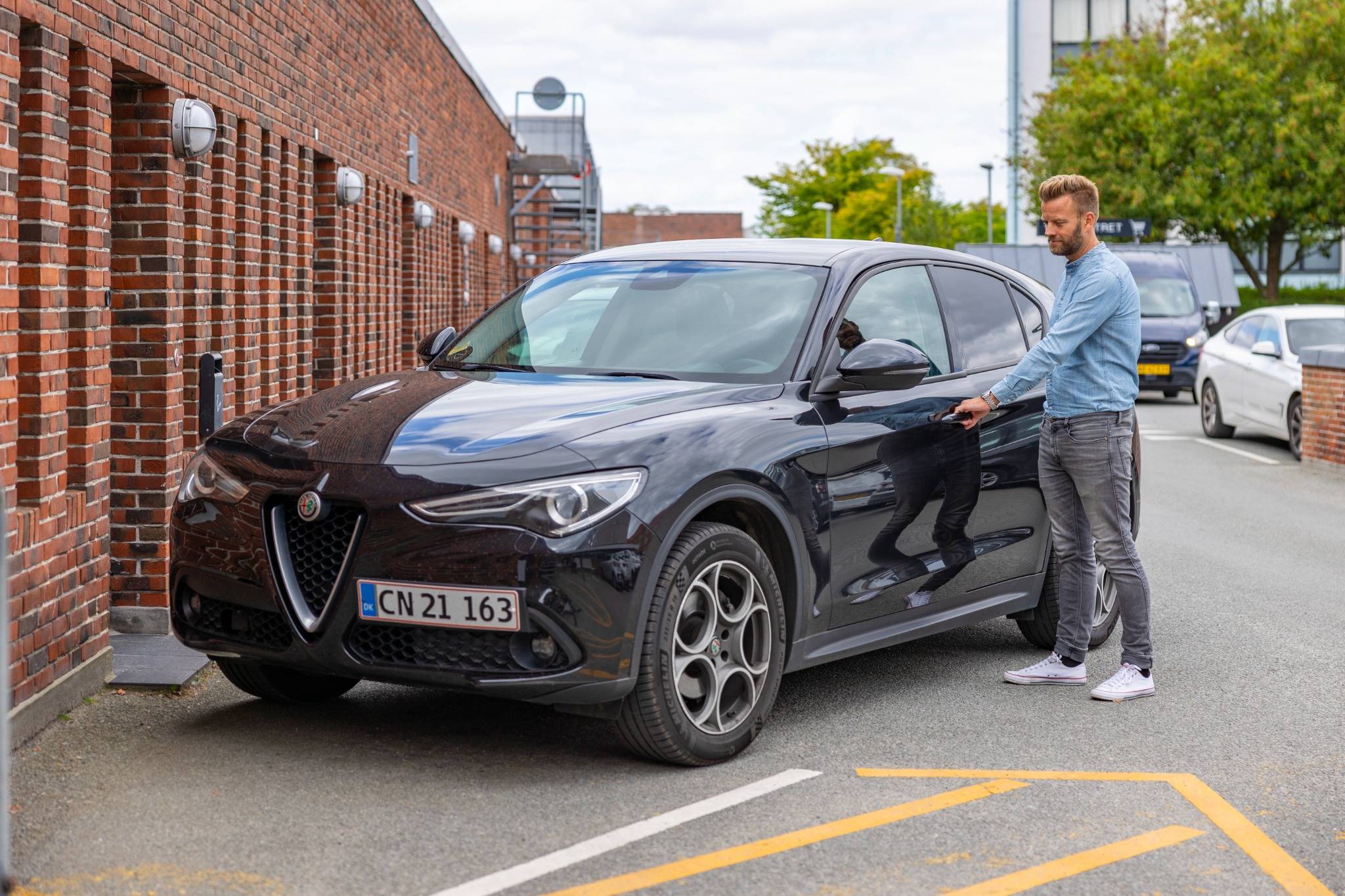 En mand iført denimskjorte og grå bukser åbner døren til en sort Alfa Romeo SUV parkeret foran en murstensbygning. Baggrunden viser træer og andre parkerede biler.
