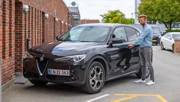En mand iført denimskjorte og grå bukser åbner døren til en sort Alfa Romeo SUV parkeret foran en murstensbygning. Baggrunden viser træer og andre parkerede biler.