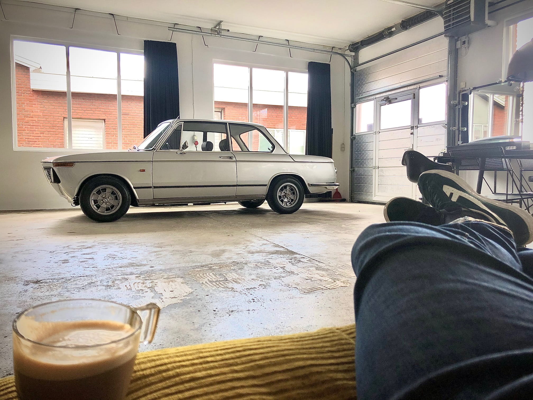 En lys garage med store vinduer og en hvid BMW 2002 klassisk bil parkeret i midten af rummet. I forgrunden ses en person, der slapper af med benene oppe på en sofa og en kop kaffe placeret på et bord foran dem. Garagen har et råt, industrielt udseende med en åben port og forskellige redskaber og møbler langs væggene.