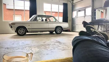 En lys garage med store vinduer og en hvid BMW 2002 klassisk bil parkeret i midten af rummet. I forgrunden ses en person, der slapper af med benene oppe på en sofa og en kop kaffe placeret på et bord foran dem. Garagen har et råt, industrielt udseende med en åben port og forskellige redskaber og møbler langs væggene.