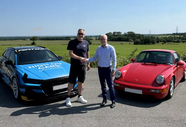To mænd står på en solrig dag på en asfalteret vej mellem to biler – en blå bil med skriften "HIGH ON CARS" og en rød klassisk Porsche. De giver hinanden et håndtryk og smiler til kameraet. Bag dem ses åbne grønne marker og træer under en klar blå himmel. Den ene mand er iført en sort t-shirt og shorts, mens den anden har en lys skjorte og mørke bukser på.