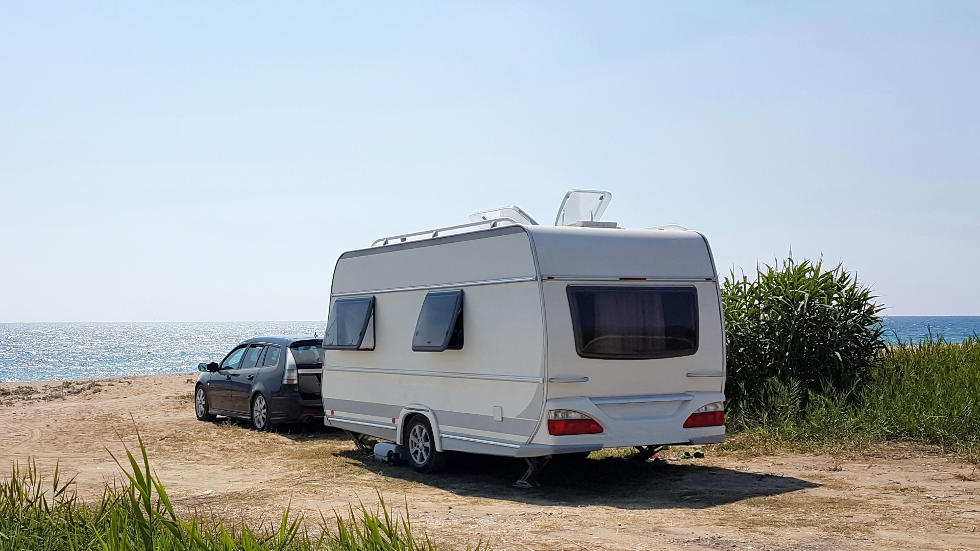 Bil med campingvogn parkeret tæt ved en strand, med udsigt til havet og omgivet af høje græsser.
