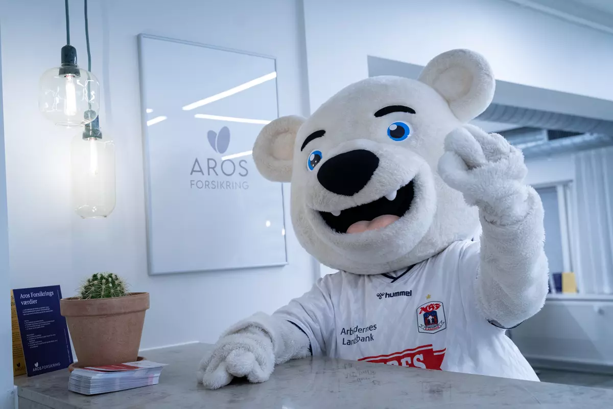 En stor, hvid bjørnemaskot iført en fodboldtrøje med Ceres-logo står bag en reception og vinker venligt mod kameraet. På væggen bag maskotten hænger et skilt med teksten AROS FORSIKRING og et logo af et blad. Til venstre på bordet står en lille kaktus i en lerpotte samt brochurer og dokumenter. Loftet har moderne industrirør og belysning.