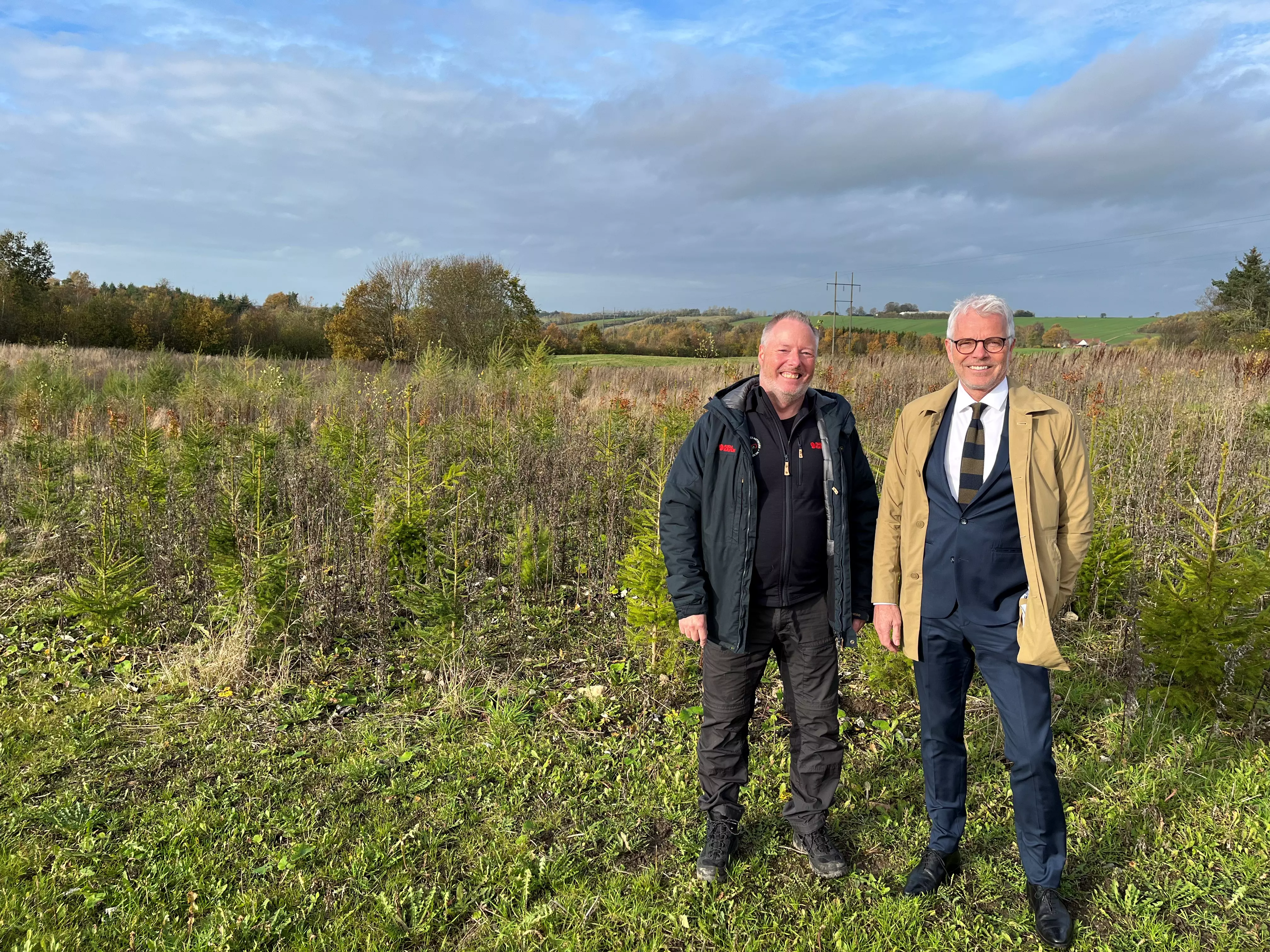 Billedet viser to mænd stående i et åbent naturområde med små træer og buske. Den ene mand til venstre har en afslappet påklædning med en mørk jakke, mens manden til højre er formelt klædt i jakkesæt og beige frakke. Begge mænd smiler mod kameraet. Baggrunden indeholder et åbent landskab med marker, træer og en delvis overskyet himmel.