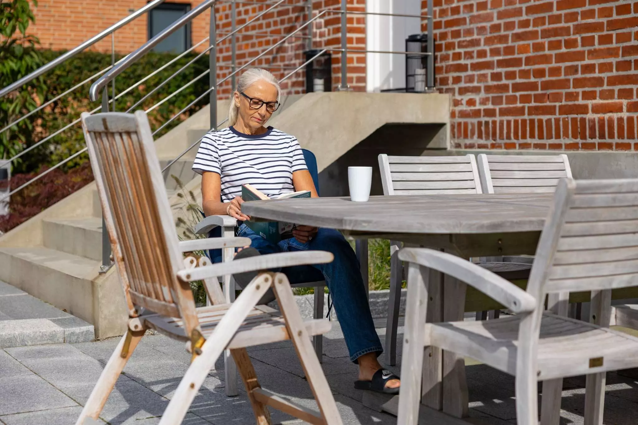 En kvinde med briller sidder udendørs på en terrasse og læser en bog. Hun har en stribet t-shirt og jeans på og sidder ved et træbord med en hvid kop. Baggrunden viser en murstensbygning og en trappe med gelænder