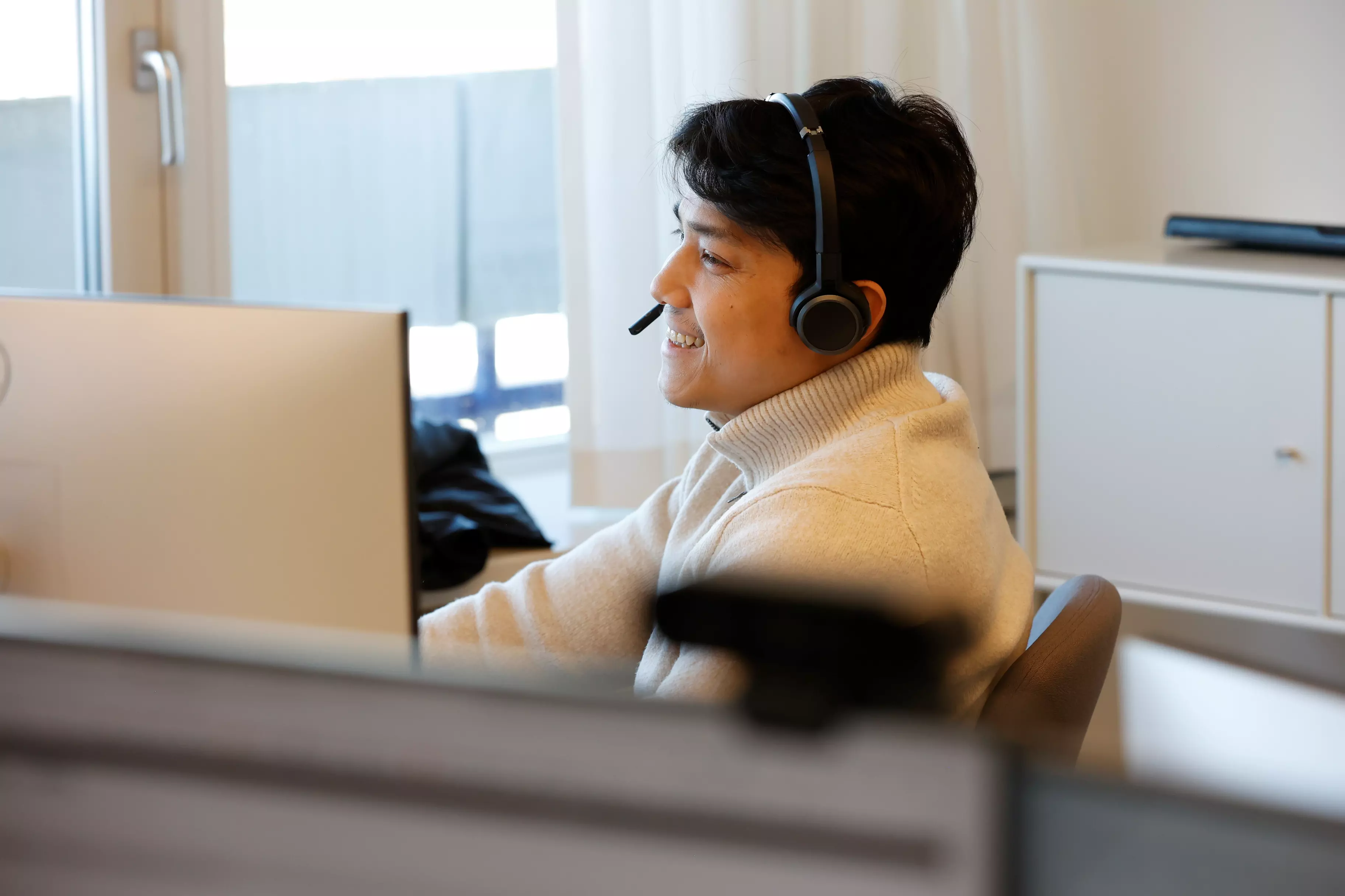 En mand sidder foran computeren i et kontormiljø. Han har headset på hovedet og smiler mens han kigger ind i computeren.