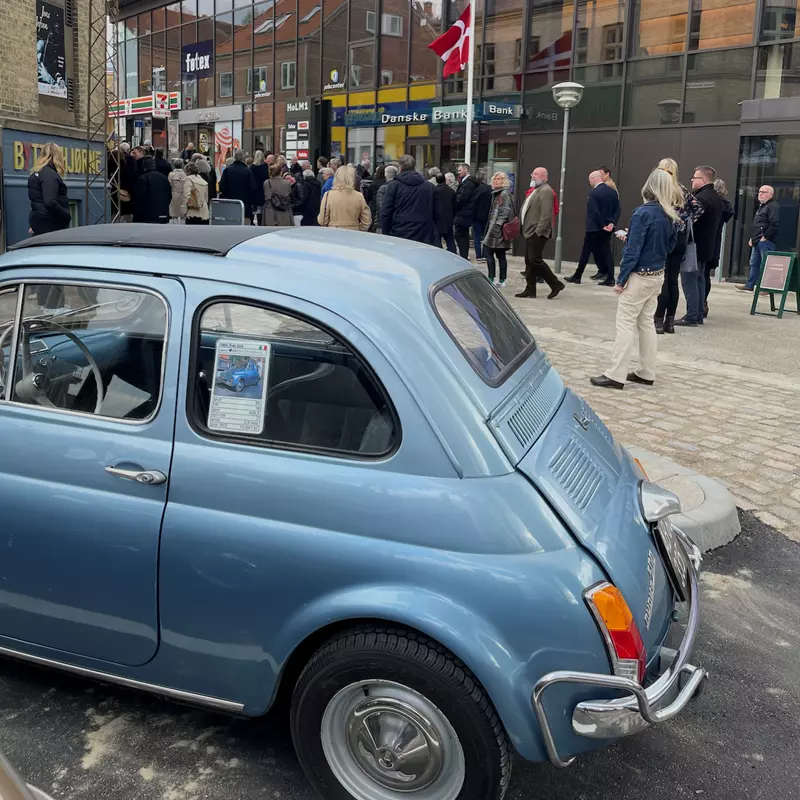 Et billede af en lyseblå vintage Fiat 500 parkeret i forgrunden på en brostensbelagt gade. Der sidder et skilt med et billede af bilen i bagruden. I baggrunden er der en gruppe mennesker i mørkt tøj, der samles foran en moderne bygning med store glasvinduer og en dansk flagstang med et flag. Flere butikker og skilte, heriblandt "Føtex" og "Danske Bank," kan ses i baggrunden.