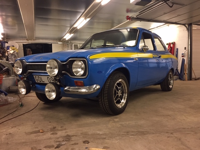 En klassisk blå Ford Escort rallybil med gule striber langs siden og store forlygter monteret foran. Bilen står i en oplyst garage med værktøj og udstyr i baggrunden, og den er tydeligvis klargjort til motorsport.