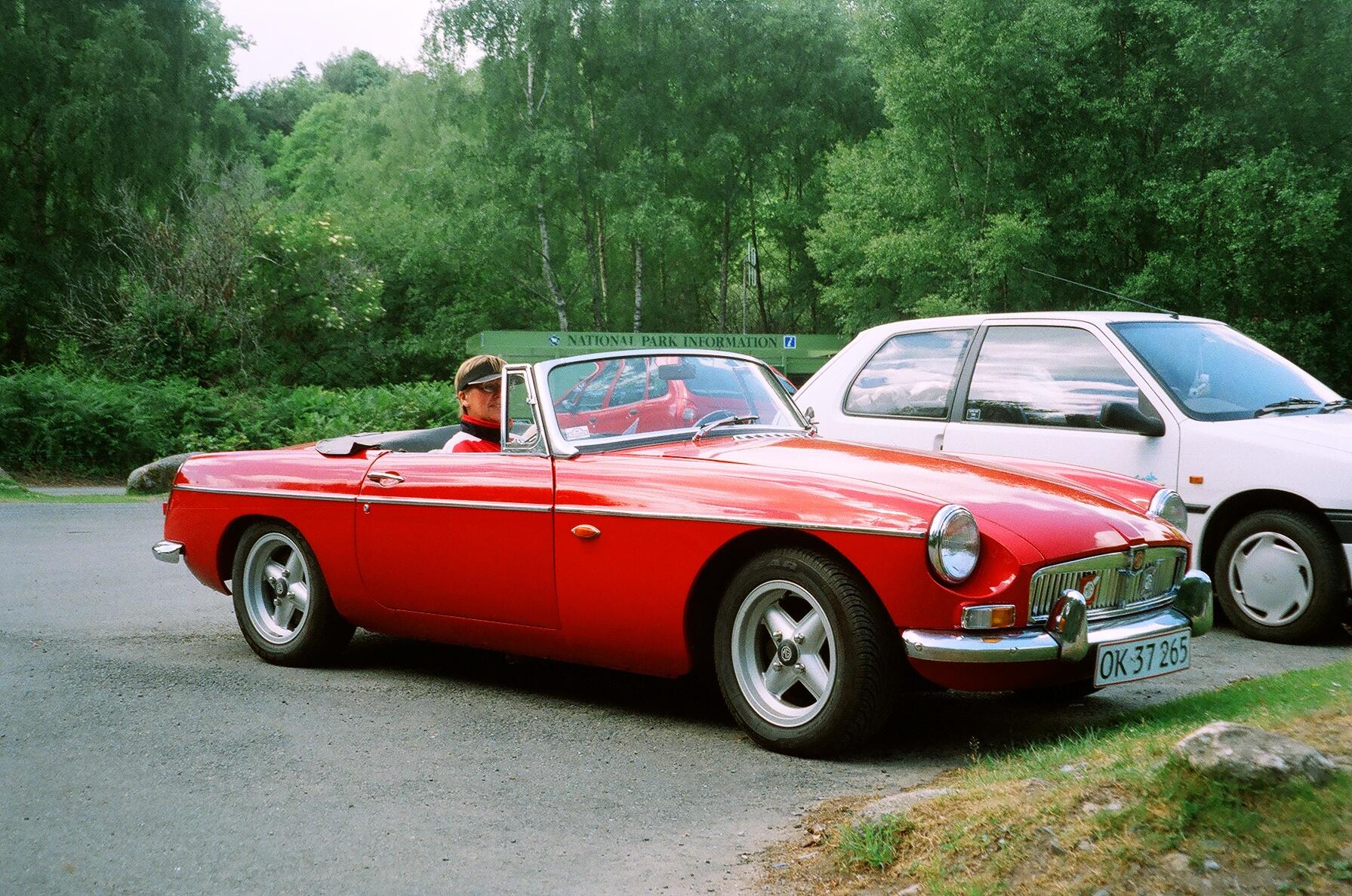 En rød klassisk sportsvogn holder parkeret i et skovområde. I bilen sidder en fører.
