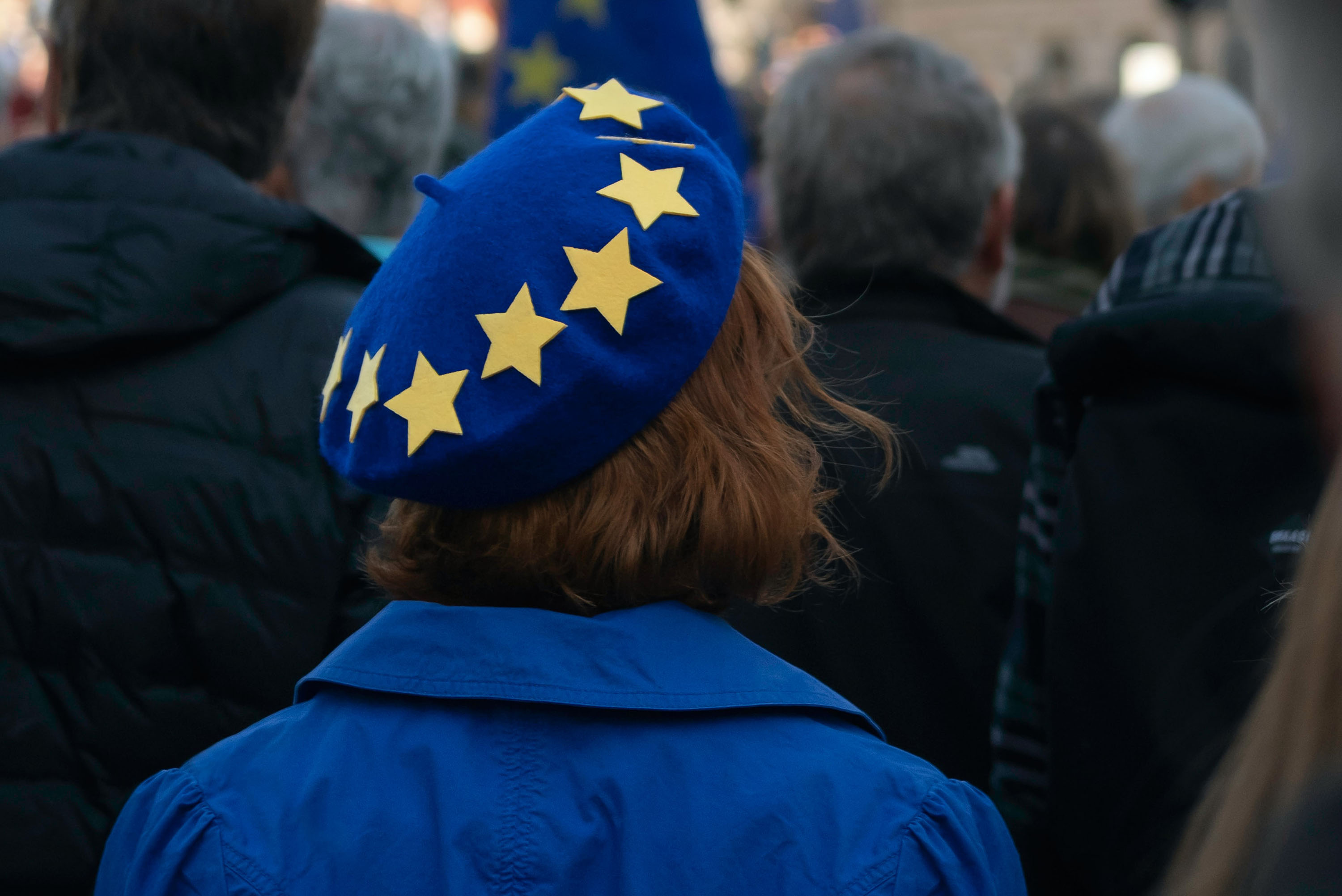 En person iført en blå baret med gule stjerner og en matchende blå jakke står blandt en folkemængde. Stjernerne på hatten ligner EU-flagets design, og baggrunden viser slørede personer i mørkt tøj.