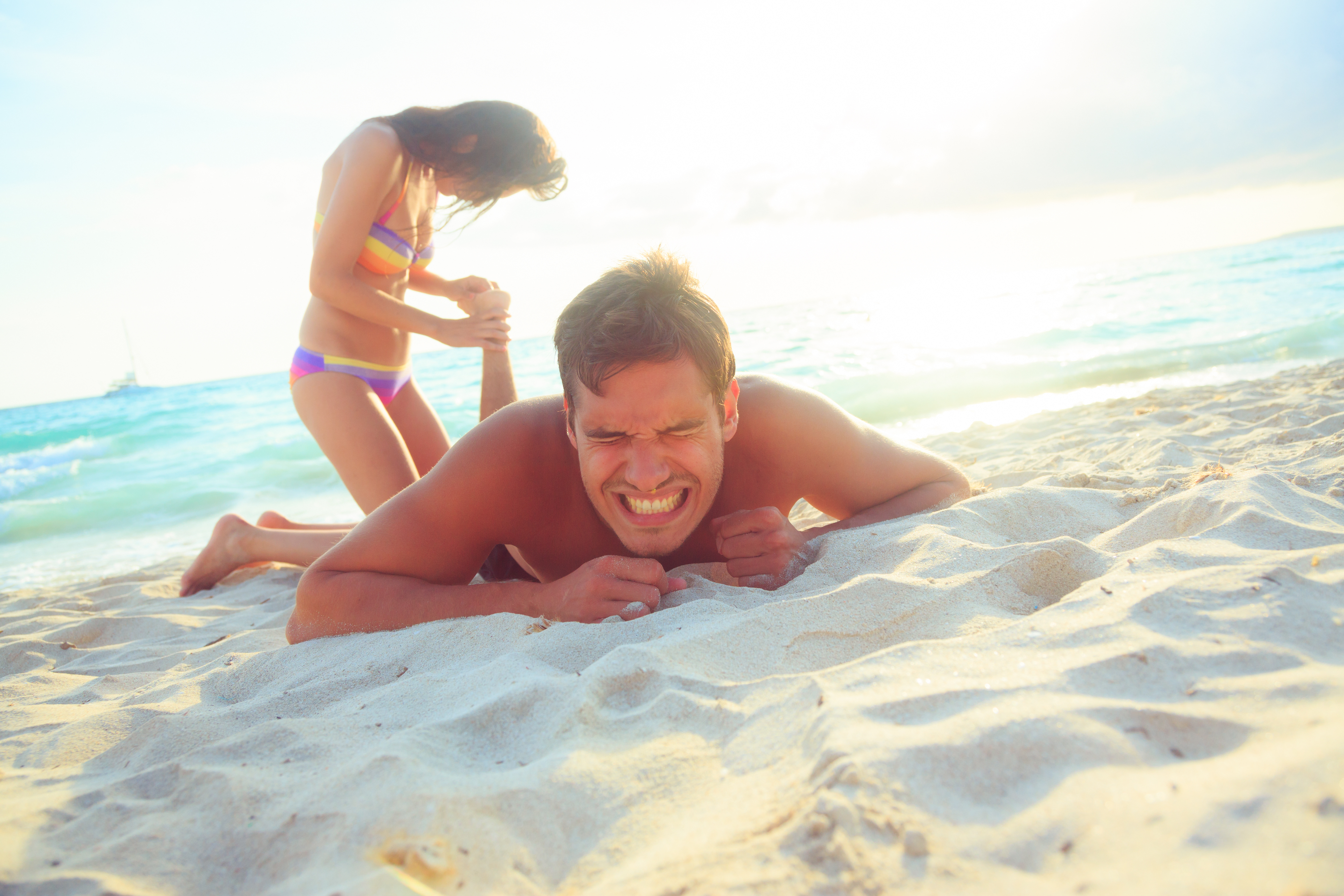 En mand ligger på en sandstrand og ser ud til at reagere på smerte, mens en kvinde i bikini står bag ham og presser på hans ryg. Baggrunden viser bølgerne fra havet og en sejlbåd i det fjerne under en solrig himmel.