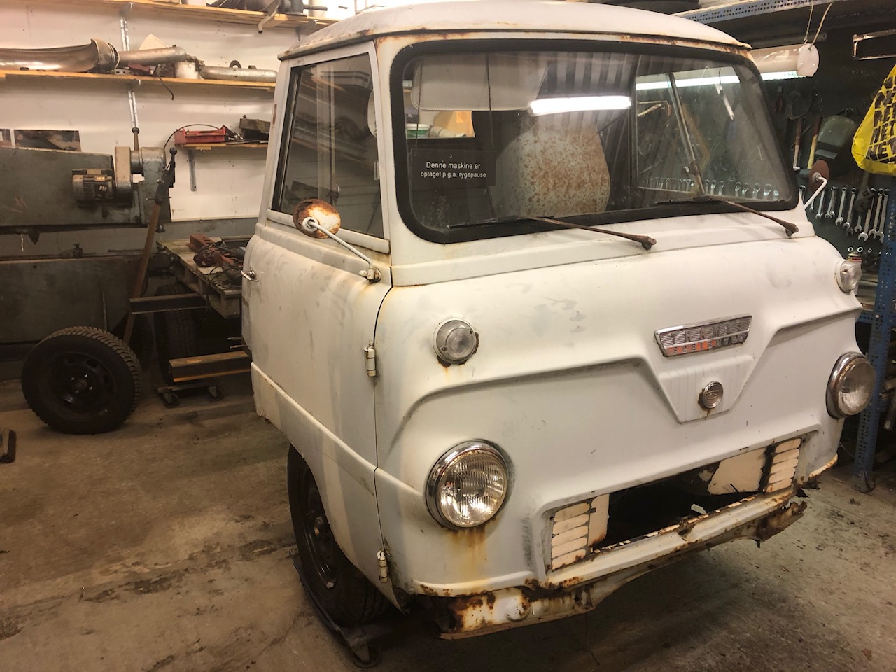 En gammel, rusten varevogn står i et værksted under reparation. Fronten af bilen er delvist adskilt med synlige rustpletter og manglende dele i kofangeren. I baggrunden ses værkstedsudstyr, som en arbejdsbænk, værktøj og en maskine til bearbejdning af metal. Atmosfæren i værkstedet er præget af slid og et aktivt arbejdsområde.