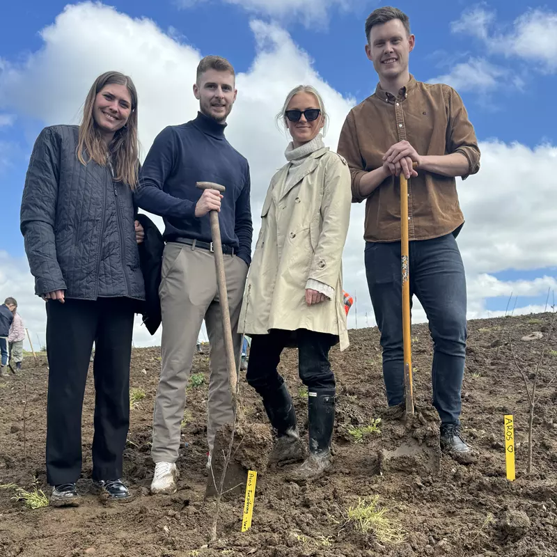 Billedet viser fire personer stående på en mudret mark med nyplantede træer eller planter i baggrunden. De er iført praktisk tøj og støvler, og to af dem holder redskaber som en skovl og et arbejdsredskab. Personerne smiler til kameraet, og der ses gule markeringer i jorden med teksten "AROS", som kunne indikere en relation til et projekt med træplantning. Himmel og natur i baggrunden understreger en udendørs aktivitet i et åbent landskab.