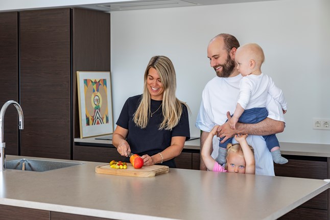 En kvinde smiler, mens hun skærer frugt på et skærebræt i et moderne køkken. Ved siden af står en mand med et spædbarn på armen, mens et lille barn kigger frem bag hans ben. Der er en farverig kunstplakat i baggrunden.