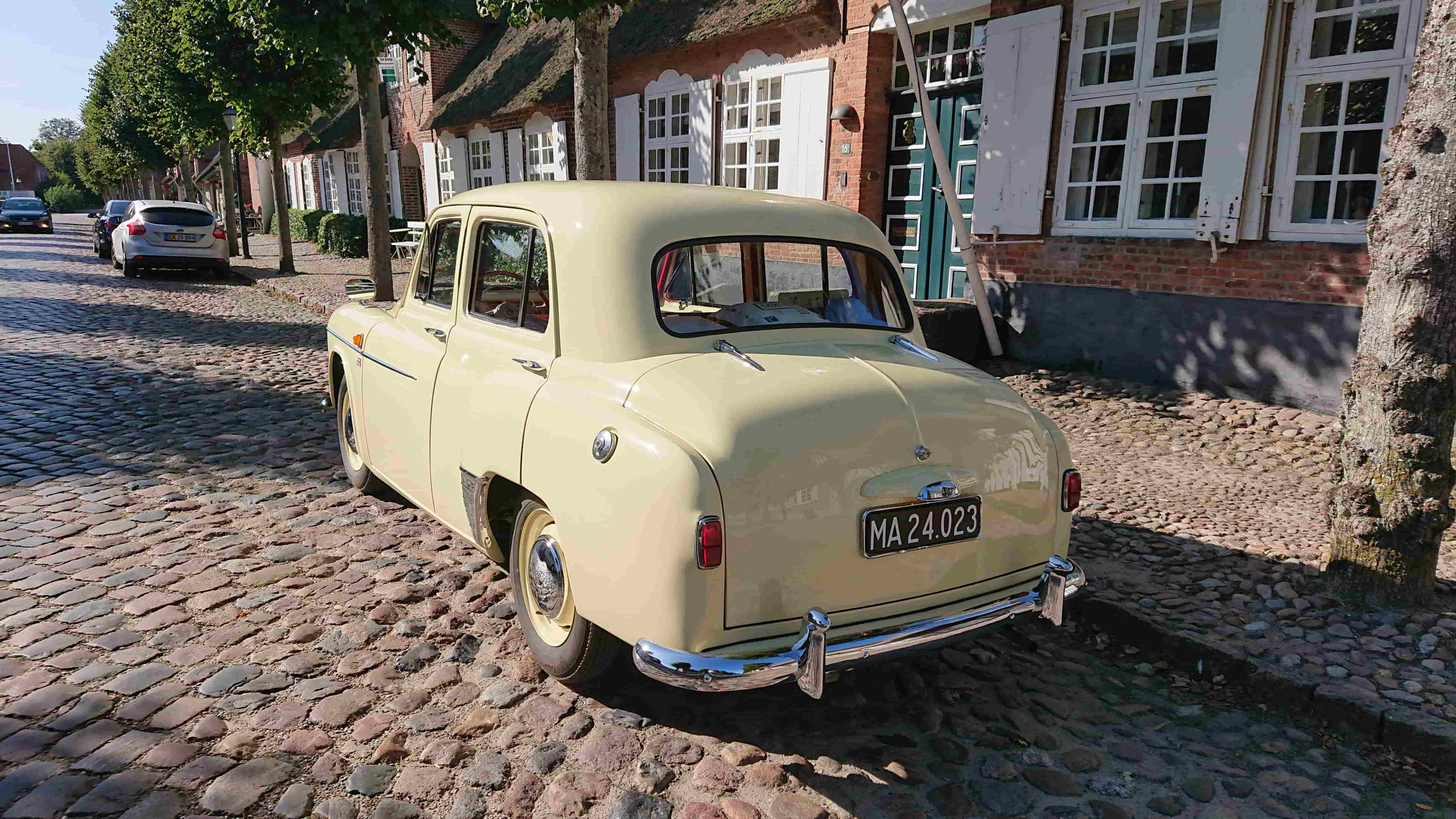 En klassisk lysegul bil parkeret på en brostensbelagt gade foran en række charmerende murstenshuse med hvide vinduer og stråtage. Solen skinner, og træer står langs vejen, hvilket skaber en hyggelig atmosfære.