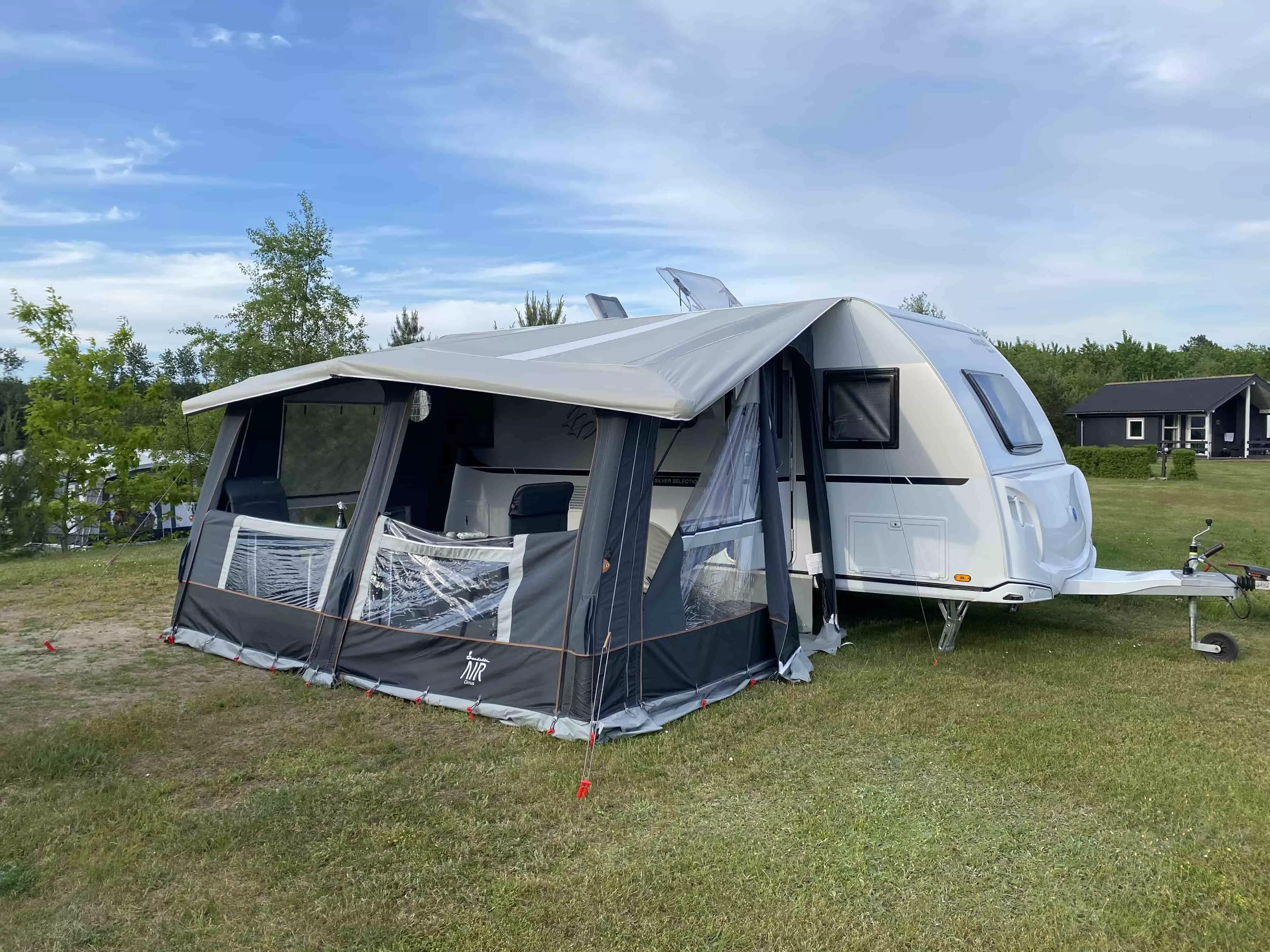 En hvid campingvogn med opslået fortelt placeret på en grøn græsplæne. Forteltet har store vinduer og er fastgjort med røde pløkker. I baggrunden ses træer og en sort træhytte.