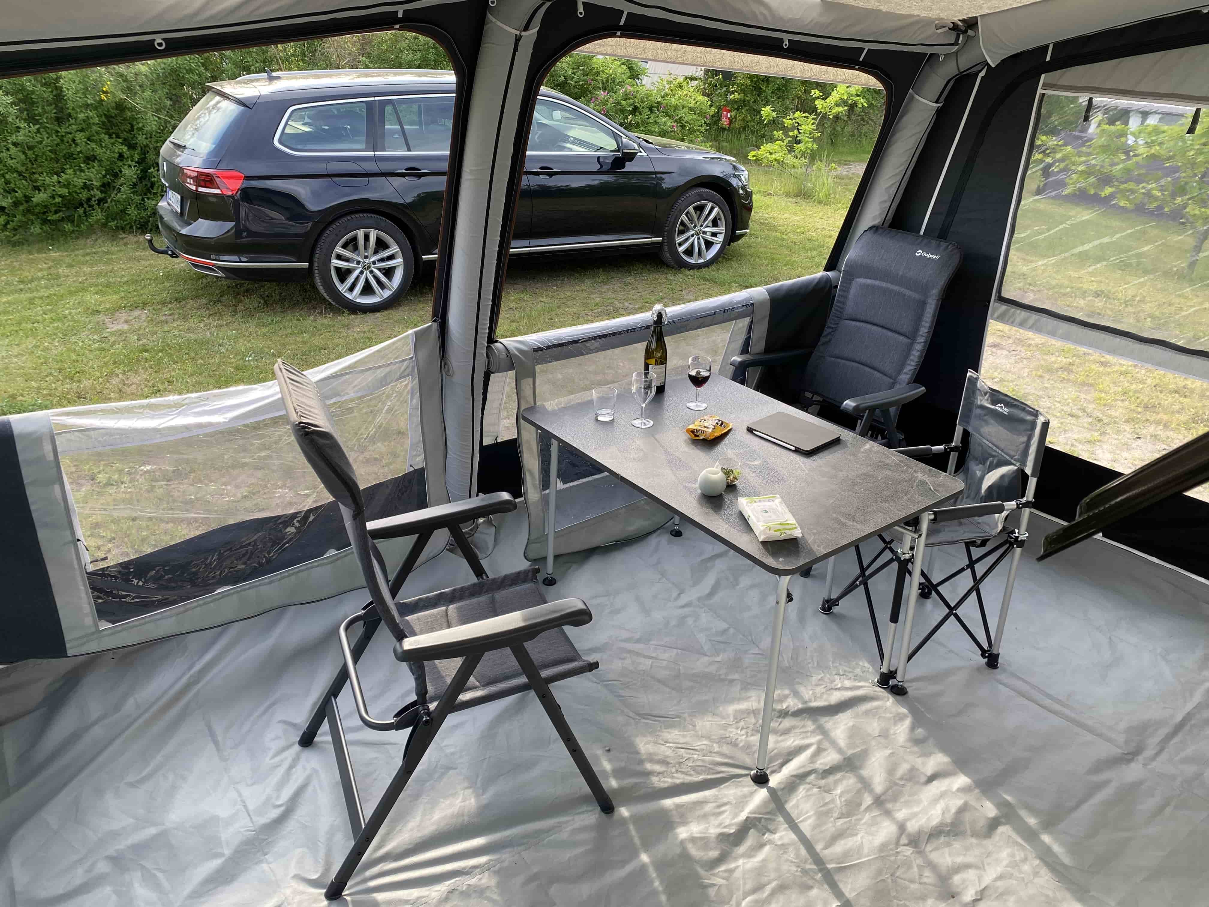 Indersiden af et campingtelt med et bord og to stole. På bordet står vinglas, en flaske vin, snacks og en mobiltelefon. Udenfor teltet holder en sort stationcar på en græsplæne omgivet af grønne buske.