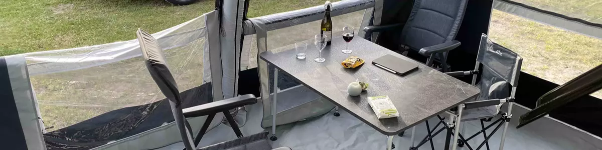 Indersiden af et campingtelt med et bord og to stole. På bordet står vinglas, en flaske vin, snacks og en mobiltelefon. Udenfor teltet holder en sort stationcar på en græsplæne omgivet af grønne buske.