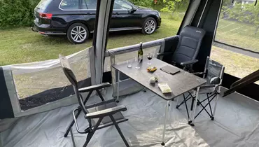 Indersiden af et campingtelt med et bord og to stole. På bordet står vinglas, en flaske vin, snacks og en mobiltelefon. Udenfor teltet holder en sort stationcar på en græsplæne omgivet af grønne buske.