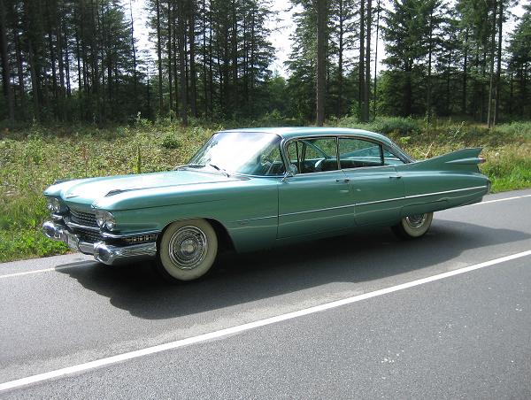 En klassisk mintgrøn Cadillac sedan fra slutningen af 1950'erne eller begyndelsen af 1960'erne, fotograferet ved en skovvej. Bilen har de karakteristiske finner bagtil, skinnende kromdetaljer og hvide dæksider, der fremhæver dens elegante og tidløse design. Med de store dimensioner og unikke linjer er den en ægte repræsentant for amerikansk bilkultur fra denne æra.