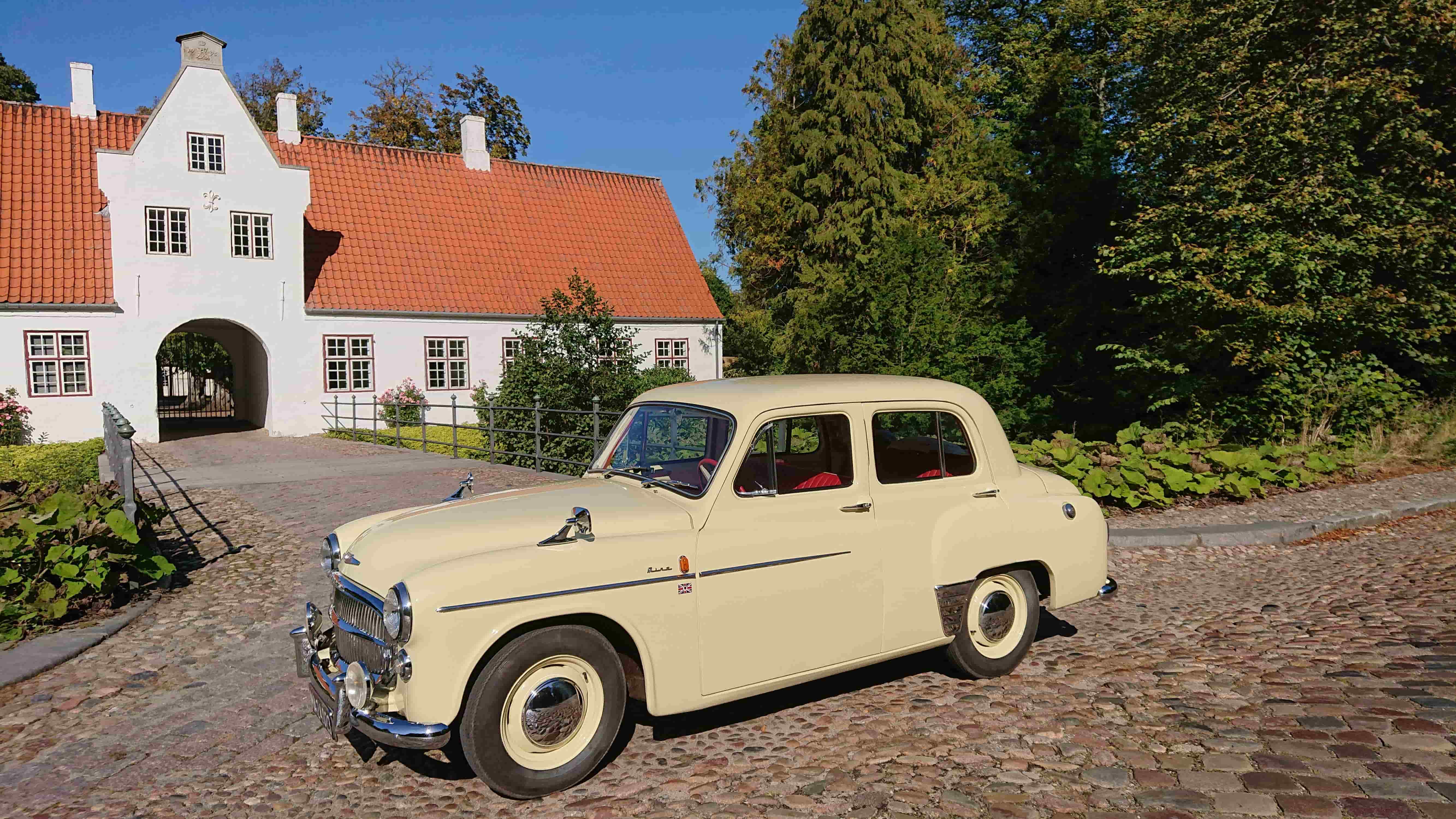En velrestaureret cremefarvet klassisk bil parkeret på en brostensbelagt vej foran en historisk hvid bygning med rødt tegltag. Bygningen har en bueformet indgang og er omgivet af frodig grøn beplantning og et klart blåt himmellandskab.