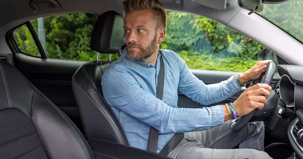 En mand iført en lyseblå denimskjorte sidder i førersædet på en bil og kigger bagud, som om han gør klar til at bakke. Bilens interiør er sort, og gennem vinduet ses grønne buske.