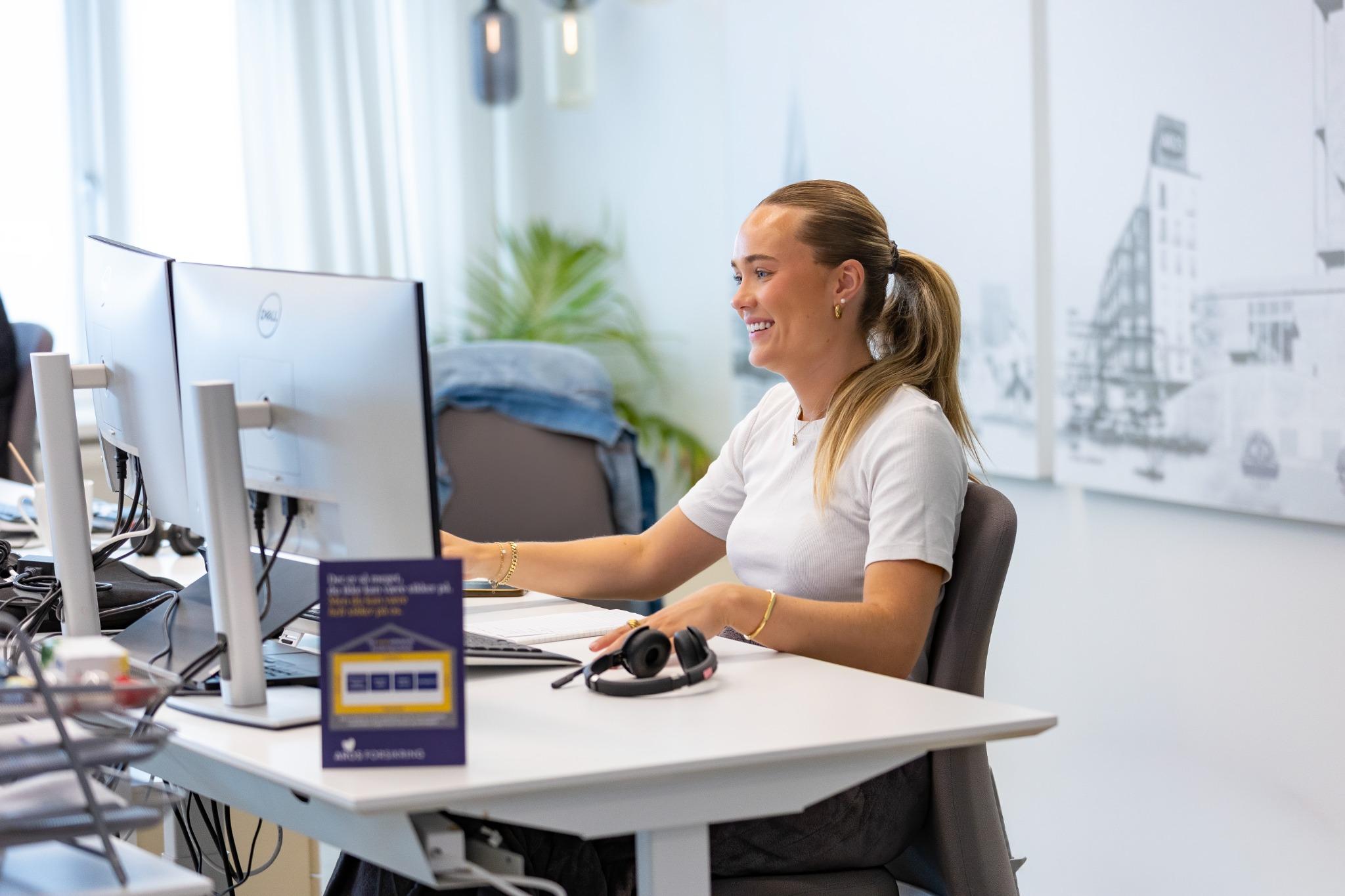 Smilende medarbejder ved hæve-sænke-bord med to skærme i lyst kontor