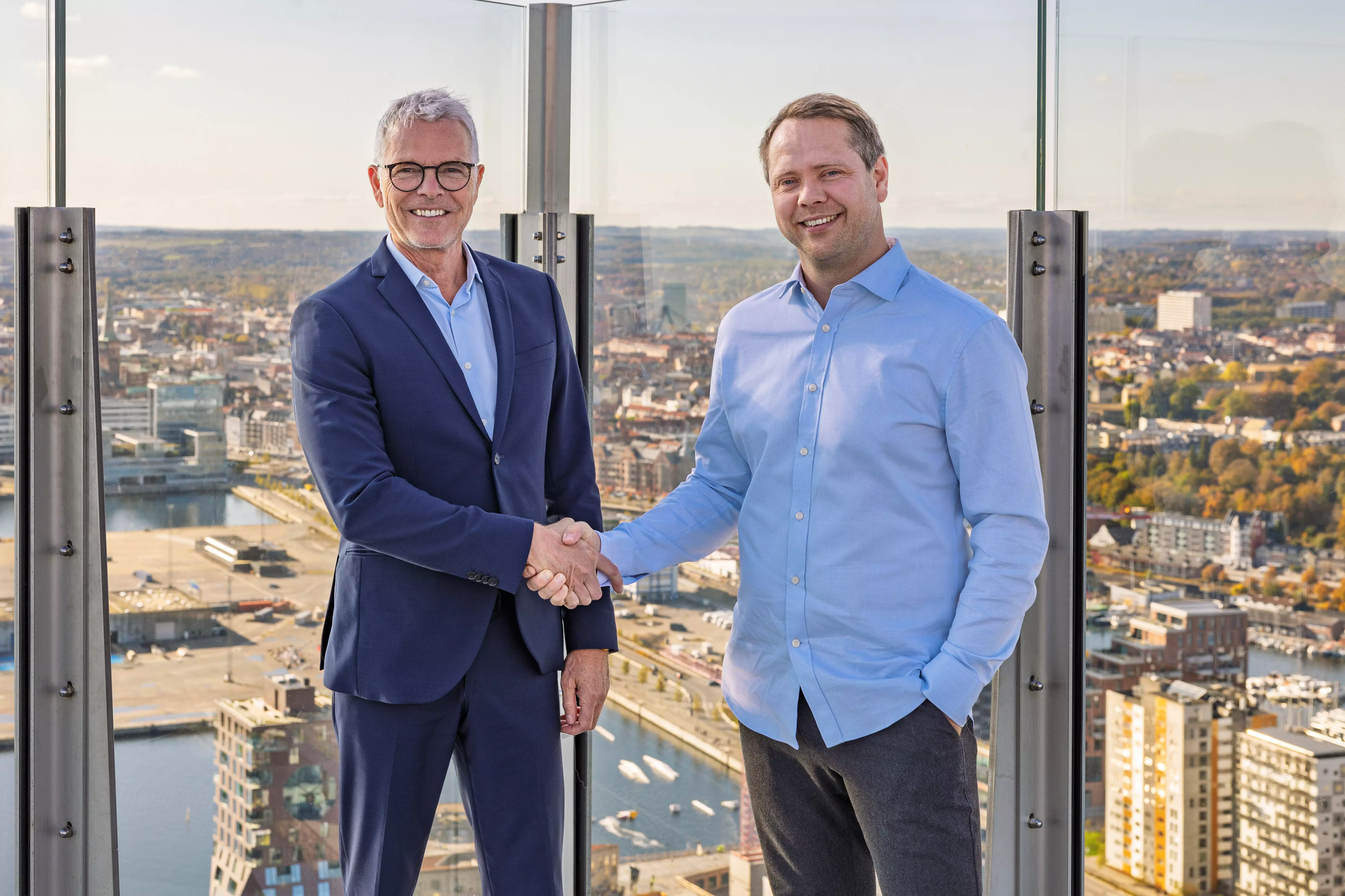 Billedet viser to mænd, der smiler og giver hinanden håndtryk på en tagterrasse med en fantastisk udsigt over en by med havn, bygninger og efterårsfarver i baggrunden. Den ene mand er iført et mørkeblåt jakkesæt med en hvid skjorte og briller, mens den anden mand bærer en lyseblå skjorte og mørke bukser. Situationen udstråler professionalisme, samarbejde og en formel, men venlig atmosfære.
