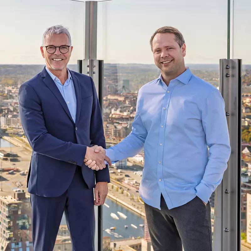 Billedet viser to mænd, der smiler og giver hinanden håndtryk på en tagterrasse med en fantastisk udsigt over en by med havn, bygninger og efterårsfarver i baggrunden. Den ene mand er iført et mørkeblåt jakkesæt med en hvid skjorte og briller, mens den anden mand bærer en lyseblå skjorte og mørke bukser. Situationen udstråler professionalisme, samarbejde og en formel, men venlig atmosfære.