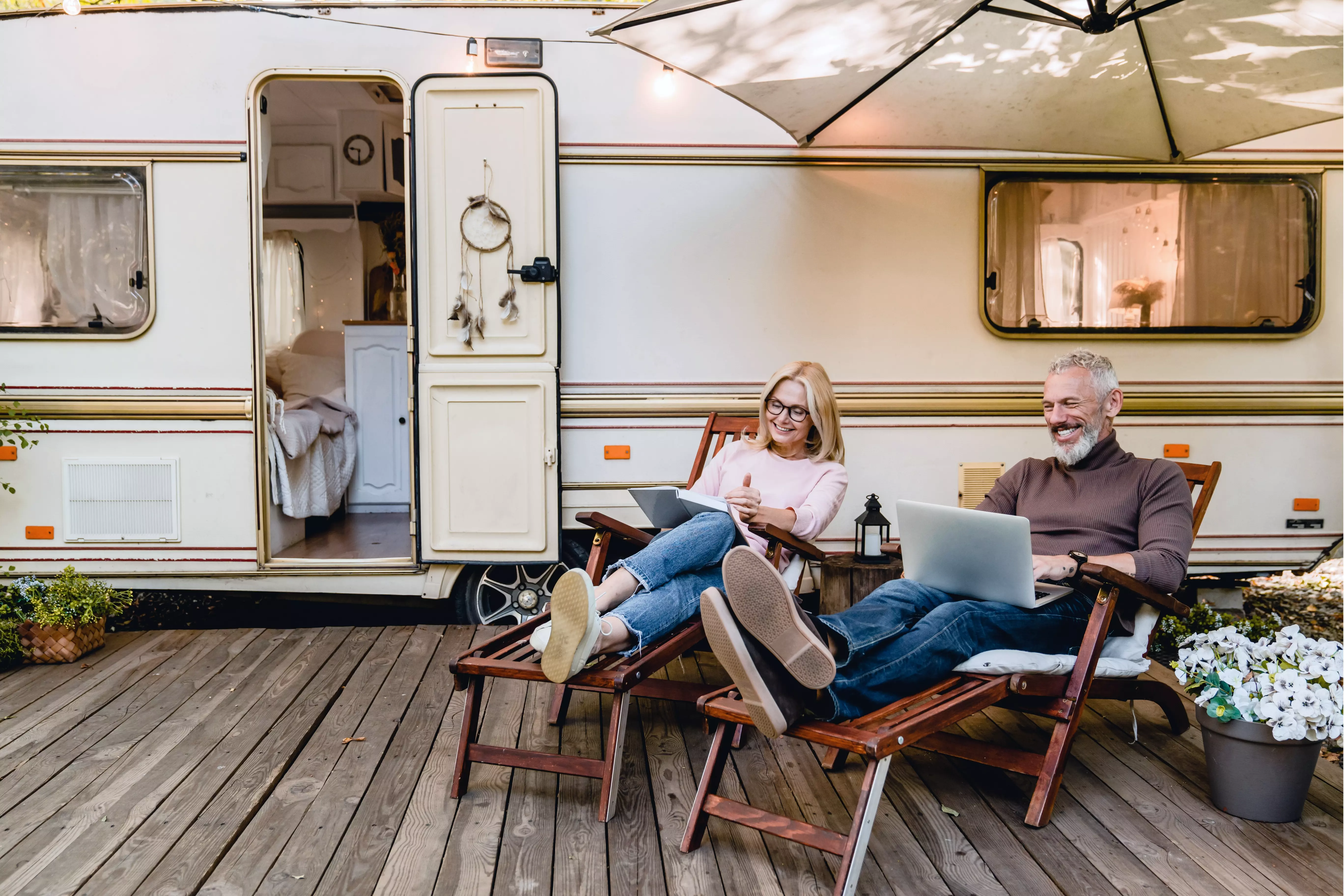 Et afslappet par sidder udendørs foran deres campingvogn på liggestole med hver deres bærbare computer. Der er et trædæk foran campingvognen, som har døren åben, så man kan se ind i det hyggelige interiør med en seng. En parasol giver skygge, og der står blomster i en krukke ved siden af campingvognen.