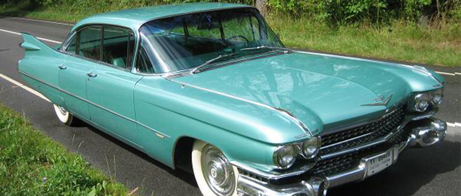En mintgrøn klassisk Cadillac fra 1950'erne eller 1960'erne, parkeret langs en asfalteret vej med grønne omgivelser. Bilen har ikoniske finner bagpå, kromdetaljer og hvide dækkanter, som fremhæver dens vintage og luksuriøse design. Dens lange, elegante linjer og skinnende lak giver den et tidløst udseende.