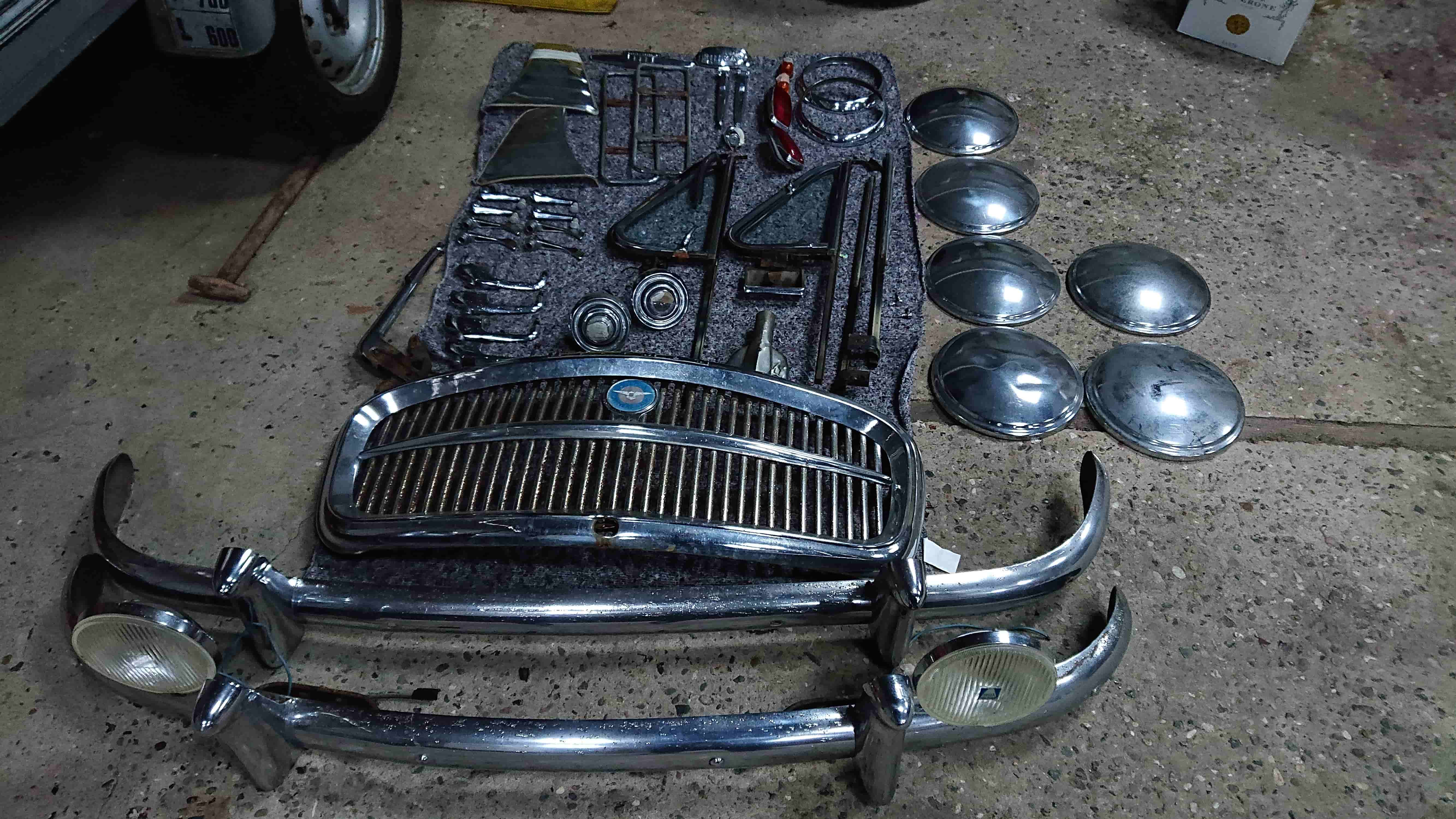 Et sæt adskilte dele fra en klassisk bil, omhyggeligt lagt på gulvet. Blandt delene ses kromgrill, kofangere med lygter, hjulkapsler og diverse beslag og trimdele. De ligger organiseret på et tæppe i en garage med betonunderlag.