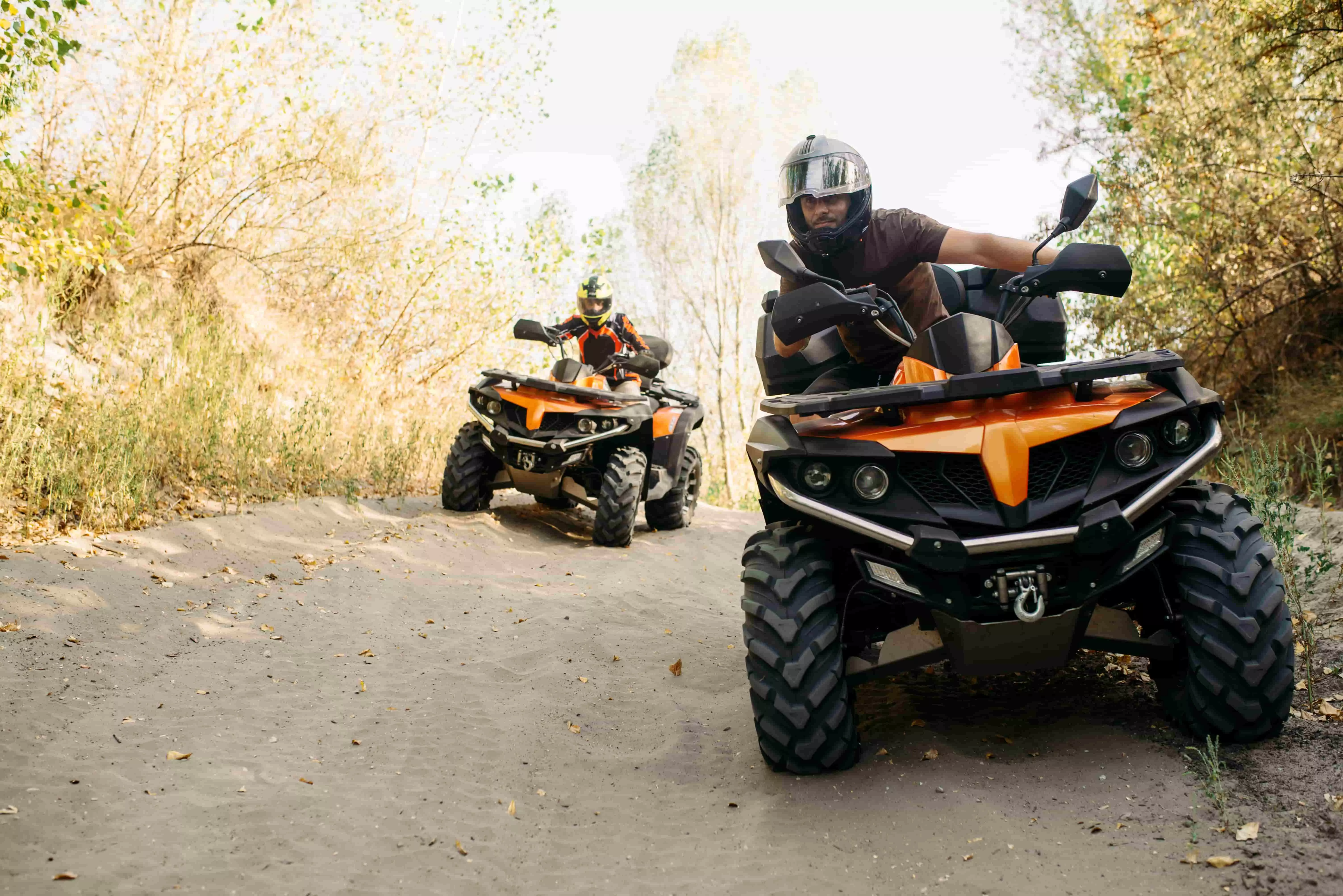 To personer på orange og sorte ATV'er kører på en støvet skovsti omgivet af tørre træer og buske, iført hjelme for sikkerhed.